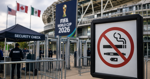 A stadium security checkpoint with a World Cup-style venue in the background and a no smoking sign in the foreground.