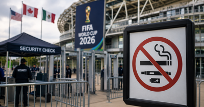 A stadium security checkpoint with a World Cup-style venue in the background and a no smoking sign in the foreground.