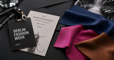 Editorial still life of a Berlin Fashion Week press pass, runway seating card, and abstract blue, pink, and brown fabric swatches on a table